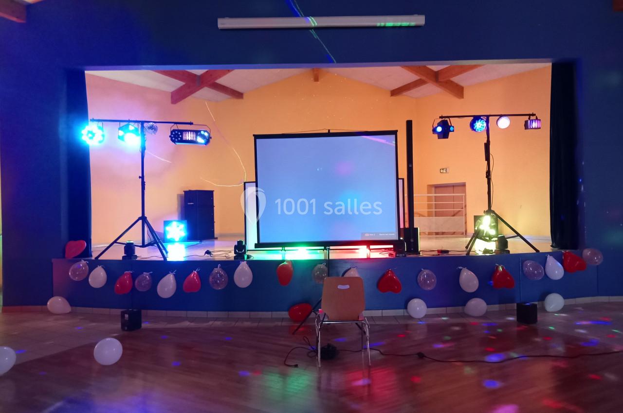 Scène décorée avec ballons, éclairages colorés, écran de projection et projecteurs dans une salle polyvalente.
