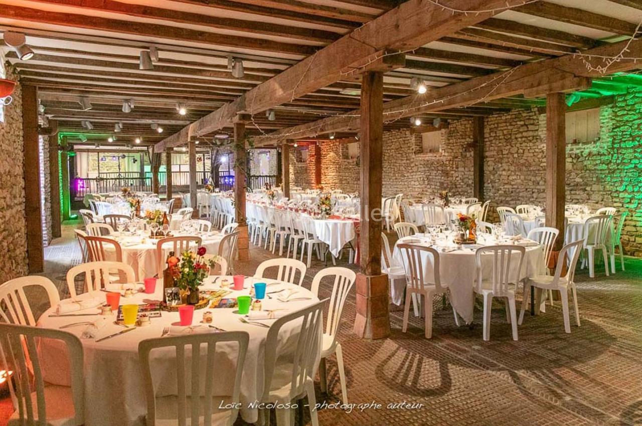 Salle de réception rustique avec tables rondes dressées, chaises blanches et décorations florales colorées.