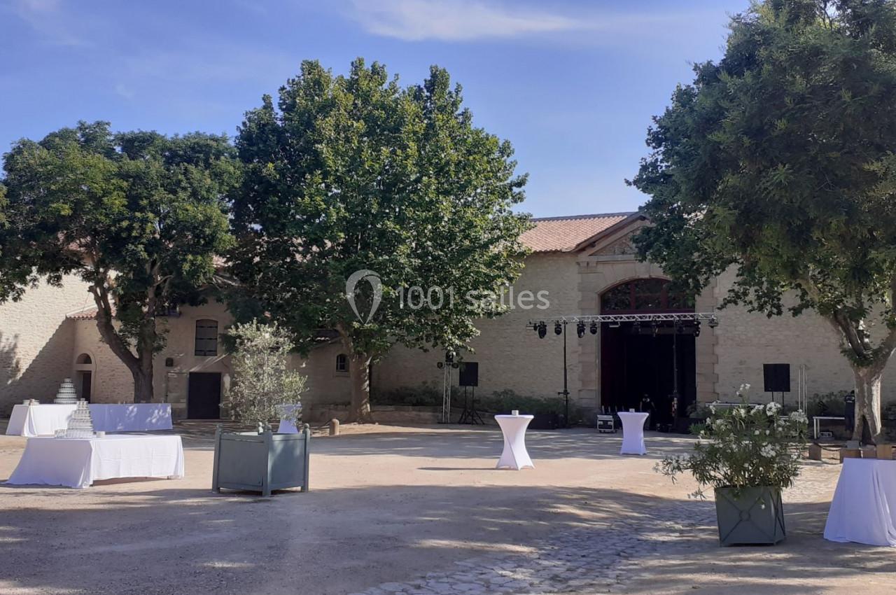 Cour extérieure avec tables hautes et nappes blanches, entourée d'arbres et de bâtiments en pierre sous un ciel dégagé.