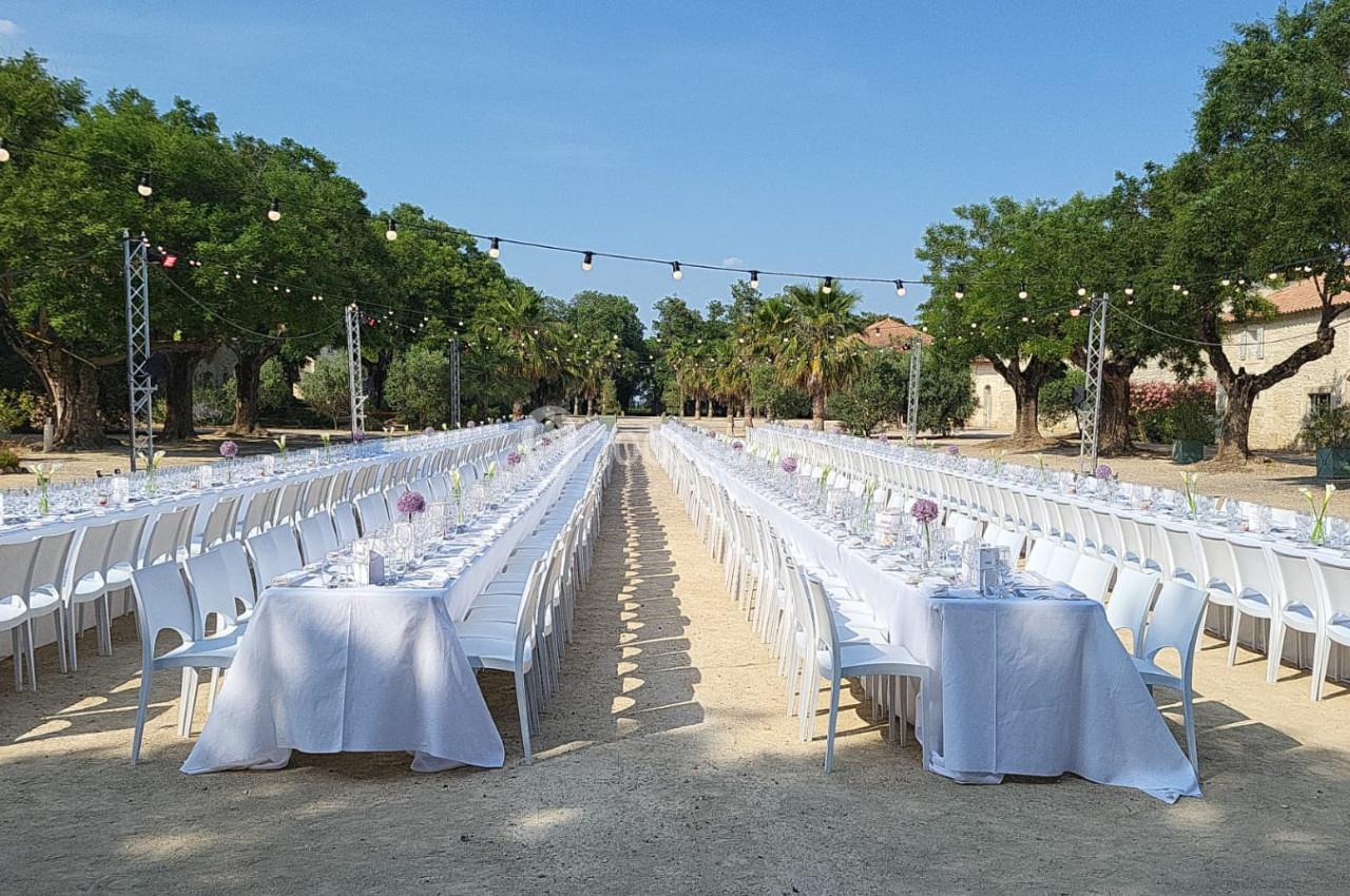 Tables longues dressées en extérieur pour un événement, entourées d'arbres et décorées de nappes blanches et de fleurs.