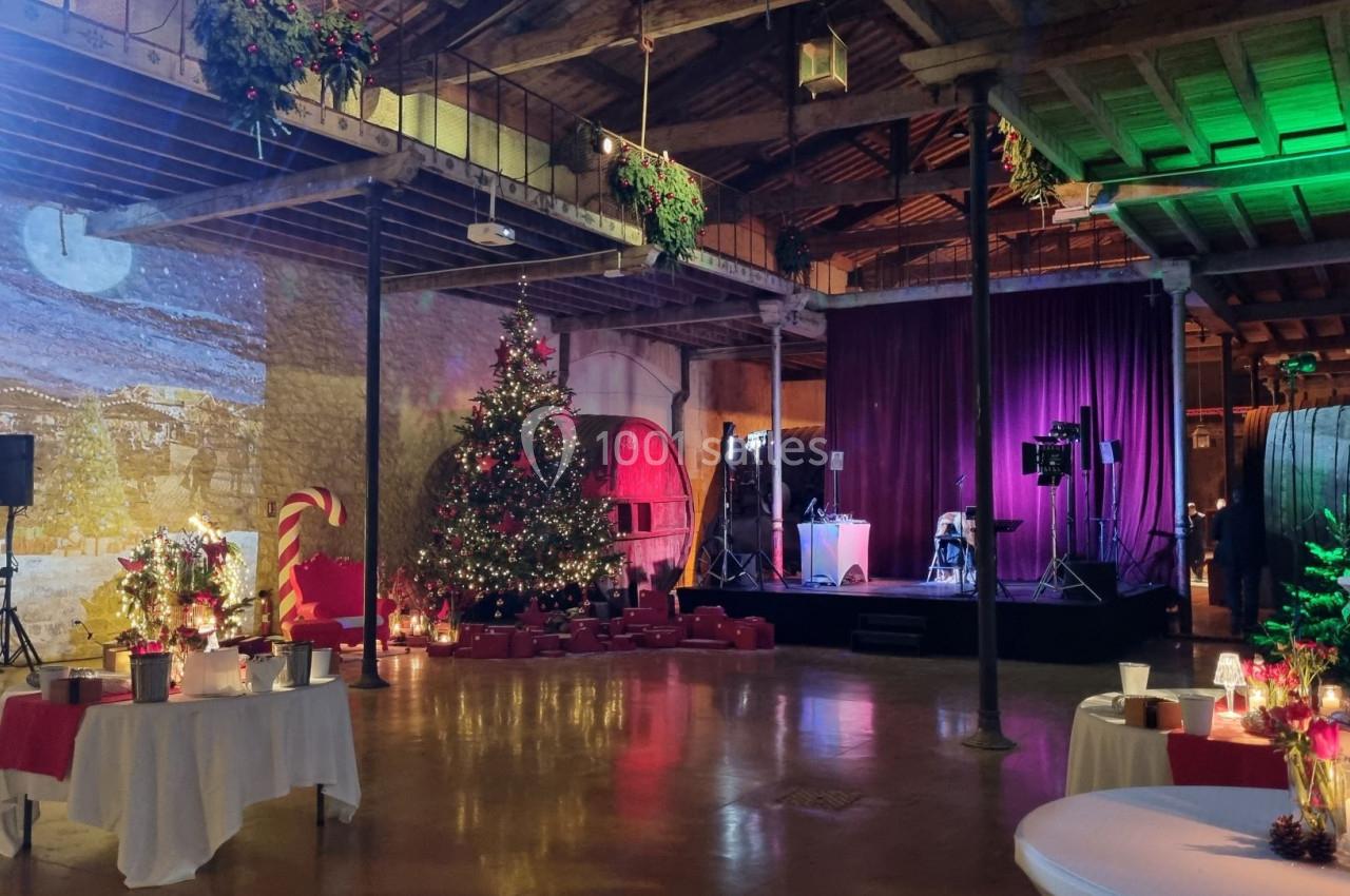 Salle décorée pour une fête avec sapin de Noël illuminé, scène avec rideaux violets et tables dressées.