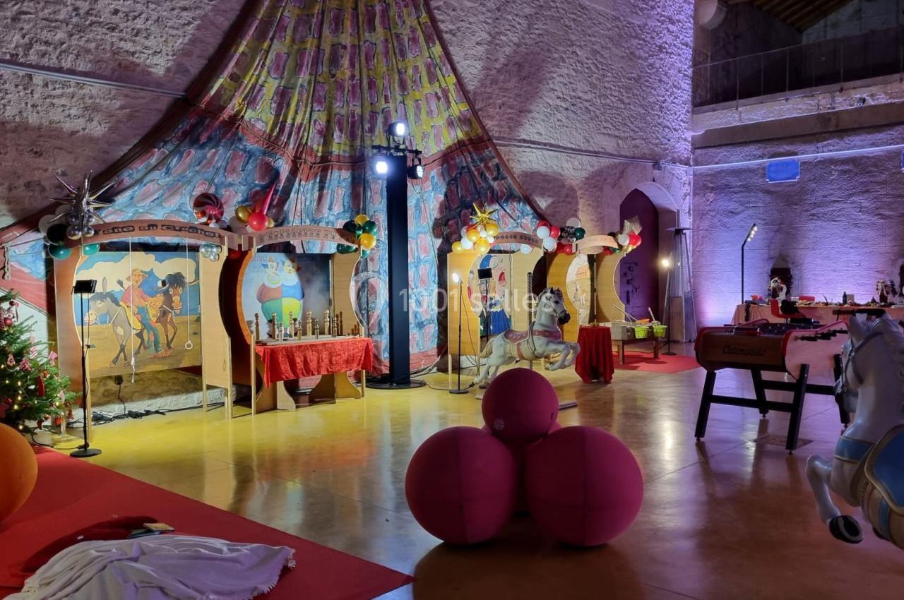 Scène intérieure avec décorations festives, chevaux de manège, ballons roses et éclairage coloré dans une salle en pierre.