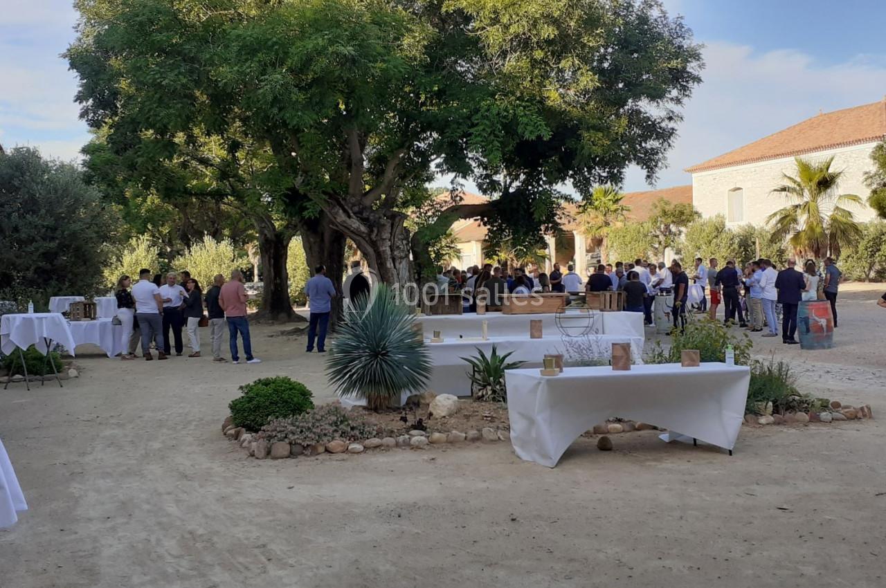 Groupe de personnes rassemblées dans une cour extérieure arborée avec des tables dressées pour un événement.