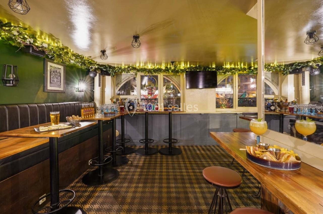 Salle de bar chaleureuse avec tables en bois, tabourets, guirlandes lumineuses et boissons disposées sur le comptoir.