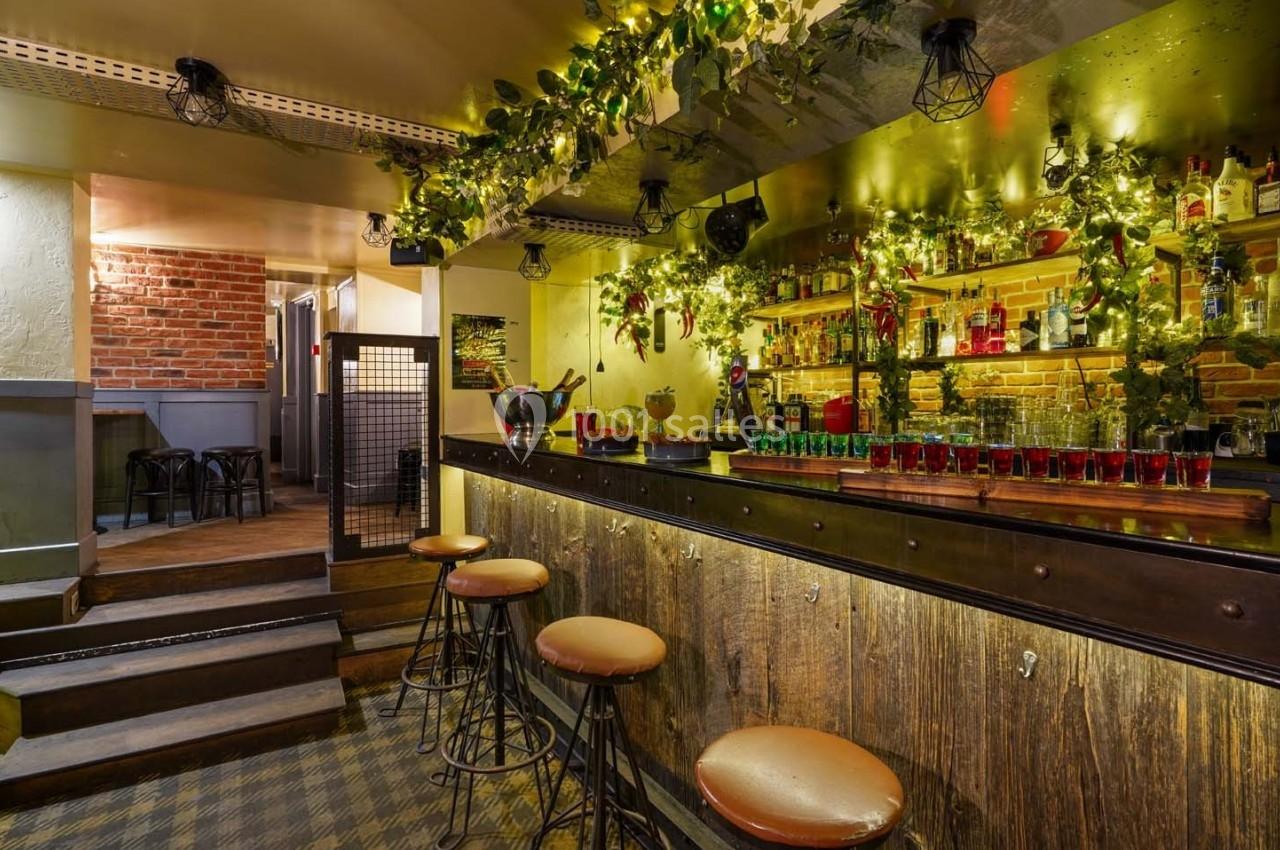 Bar chaleureux avec comptoir en bois, tabourets hauts, étagères garnies de bouteilles et décorations lumineuses végétales.