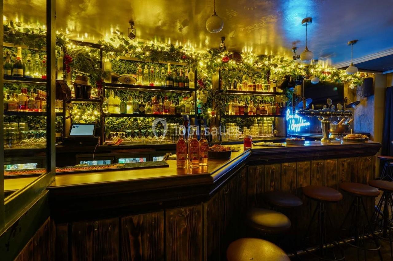Bar décoré de guirlandes lumineuses, étagères remplies de bouteilles et tabourets alignés devant un comptoir en bois.