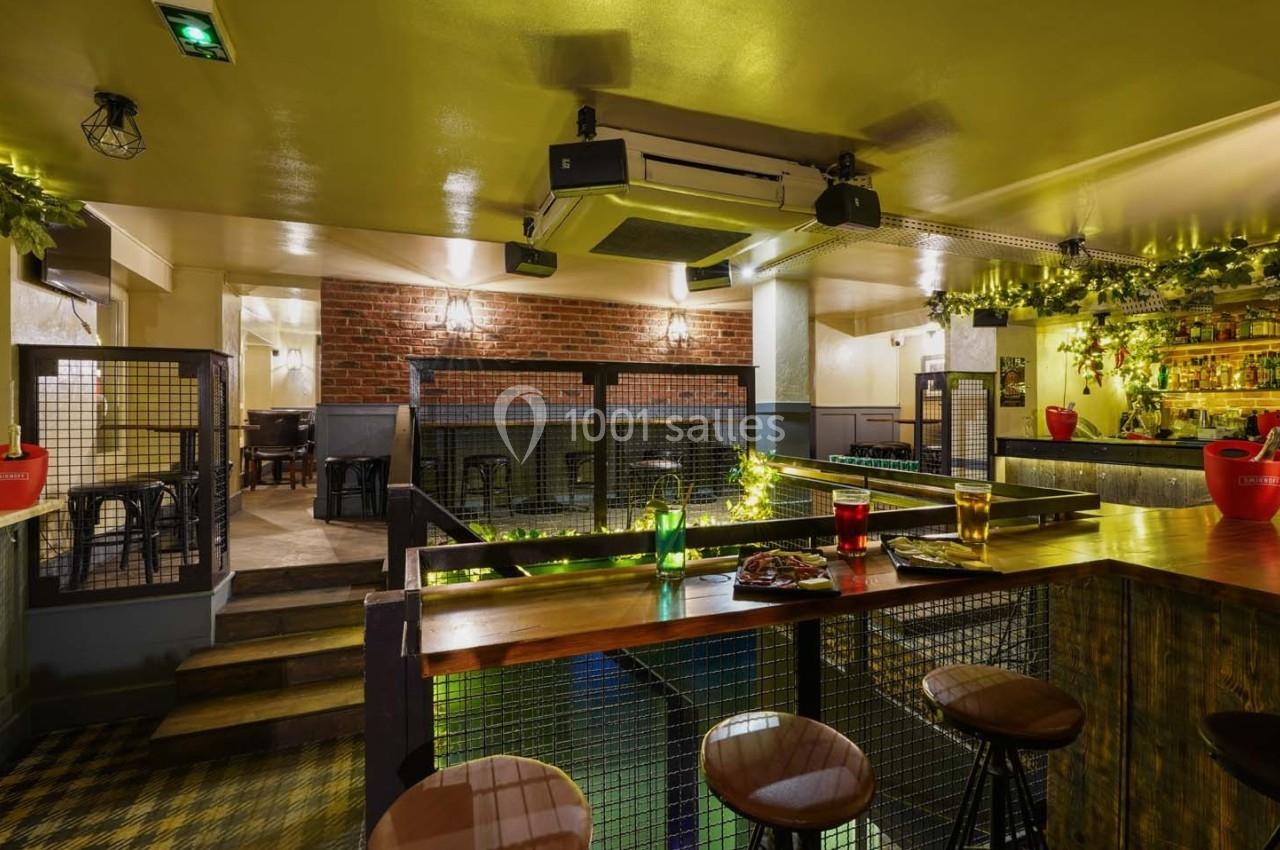 Intérieur d'un bar avec un comptoir en bois, des tabourets, un éclairage chaleureux et une décoration végétale.