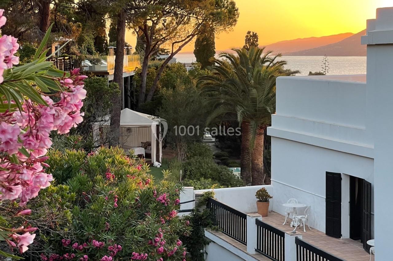 Vue sur un jardin méditerranéen avec des lauriers roses, des palmiers et une terrasse, au coucher du soleil près de la mer.