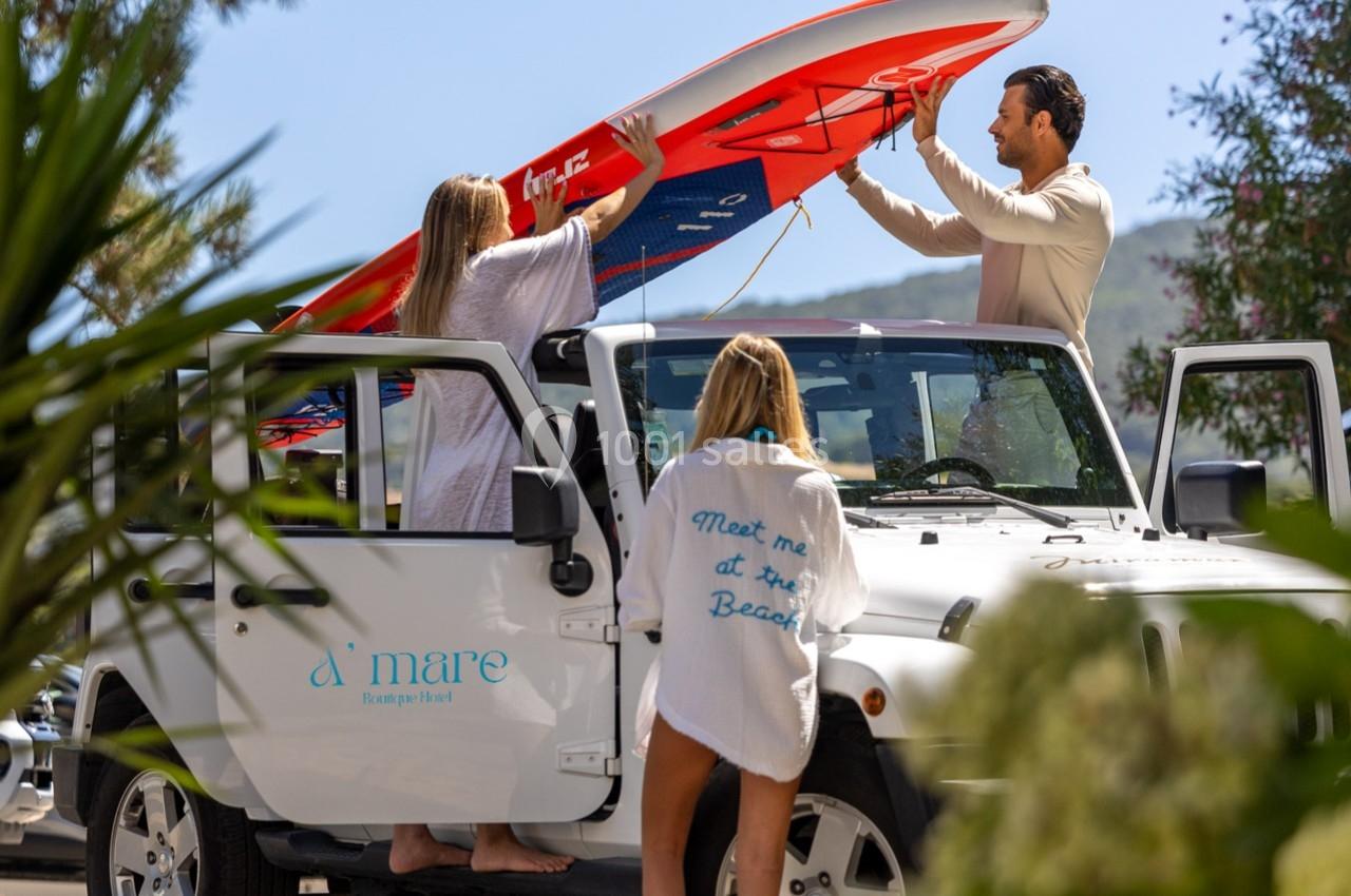 Deux femmes et un homme chargent une planche de surf rouge sur un 4x4 blanc, entourés de végétation sous un ciel bleu.