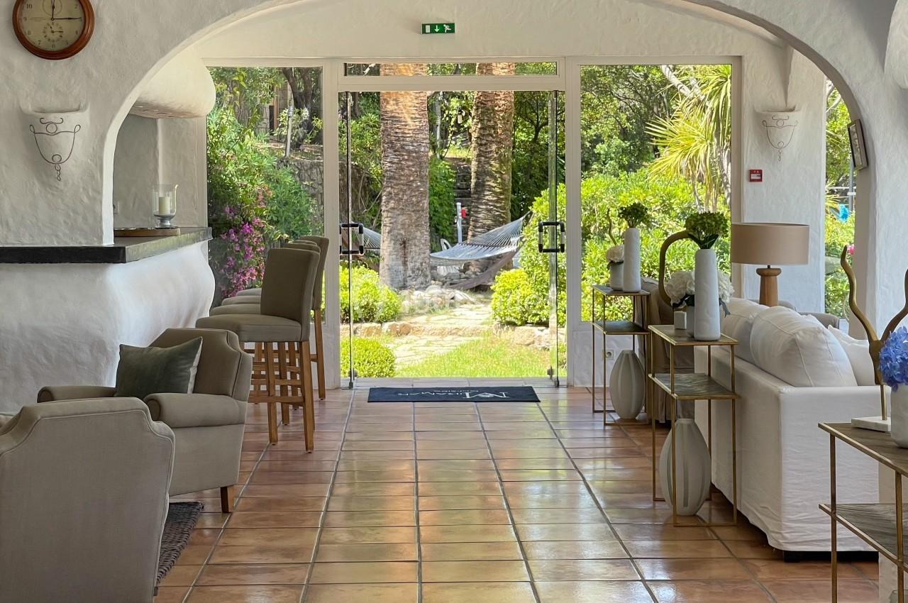 Salon lumineux avec fauteuils, bar et grandes baies vitrées donnant sur un jardin verdoyant.