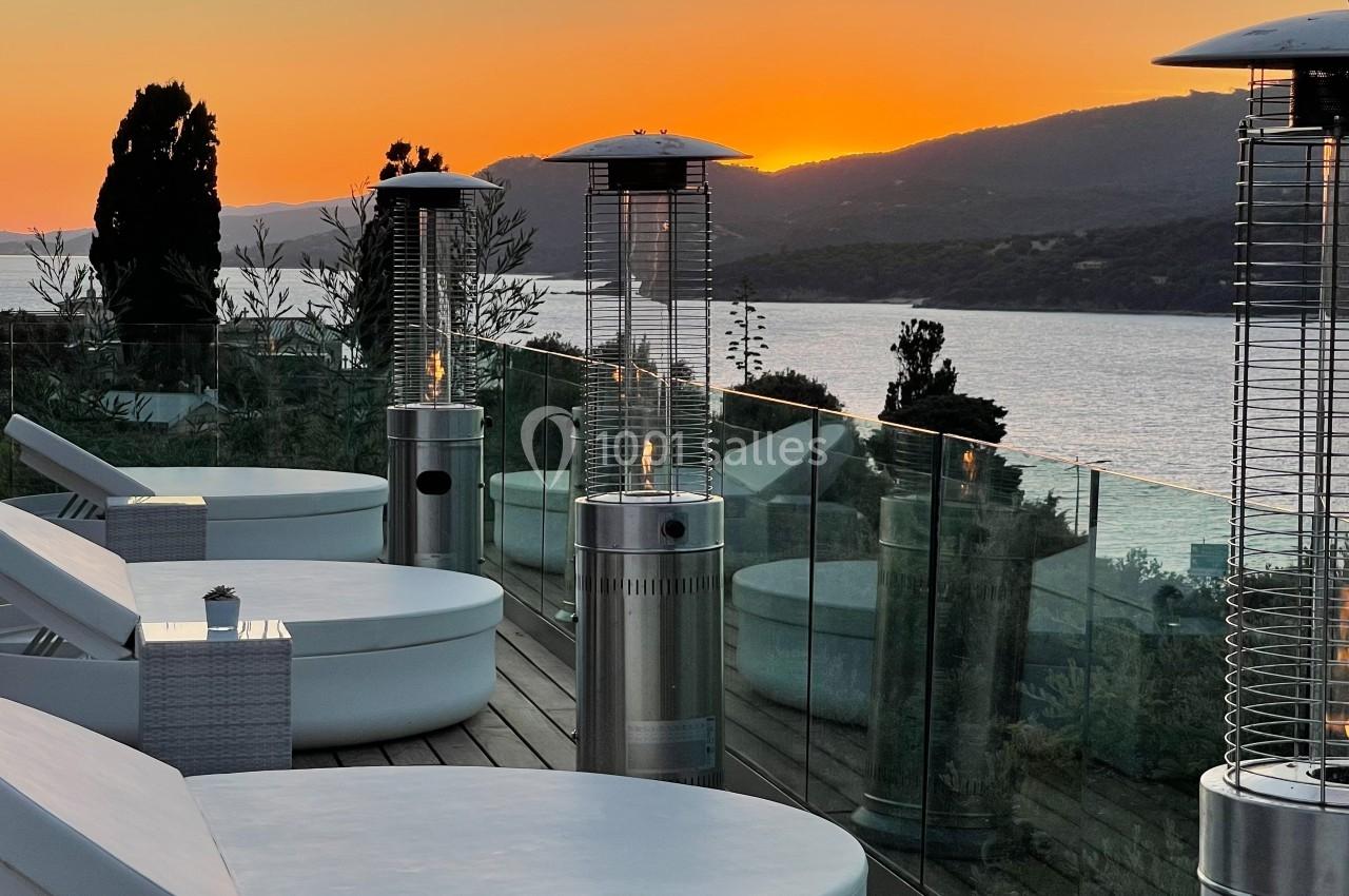 Terrasse avec chaises longues, chauffages extérieurs et vue sur un lac entouré de collines au coucher du soleil.