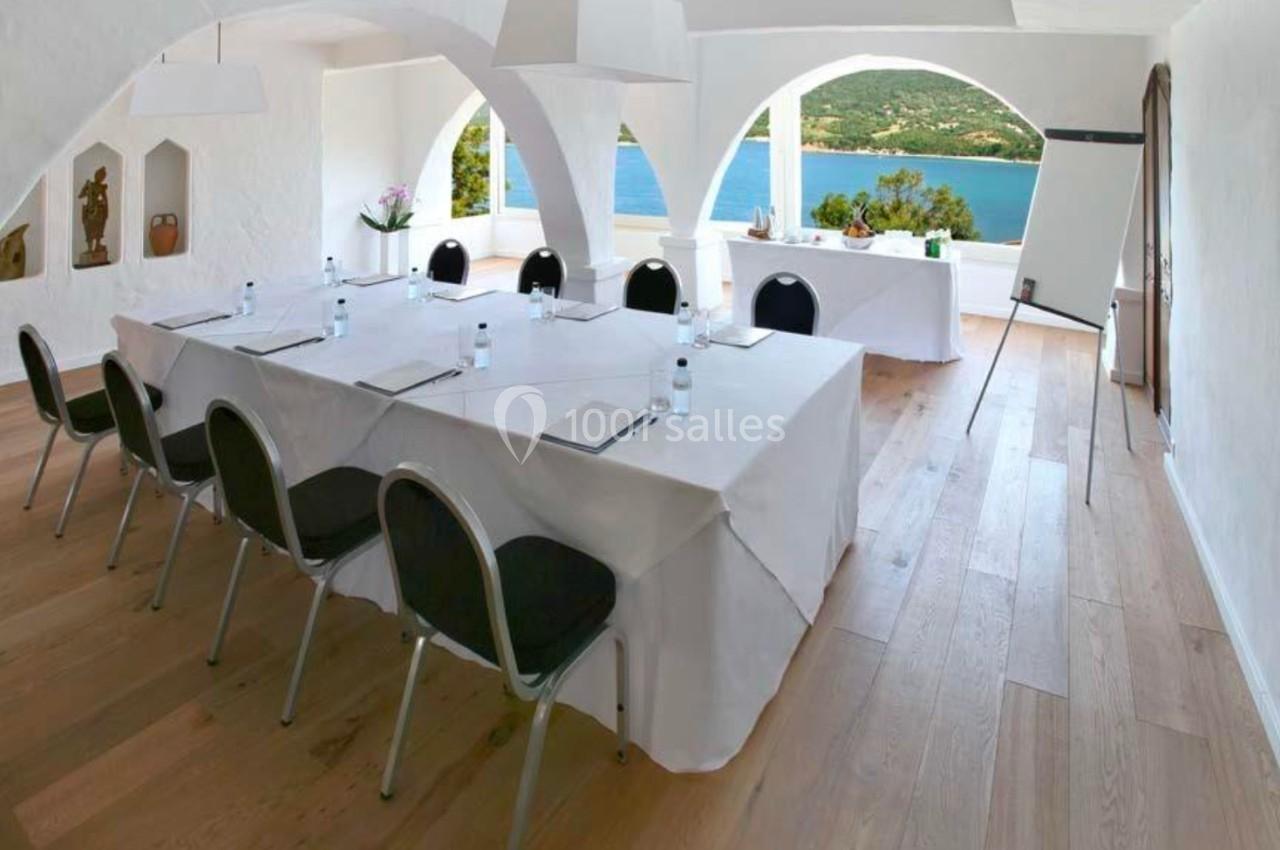 Salle de réunion lumineuse avec tables blanches, chaises noires et vue sur la mer à travers de grandes arches.
