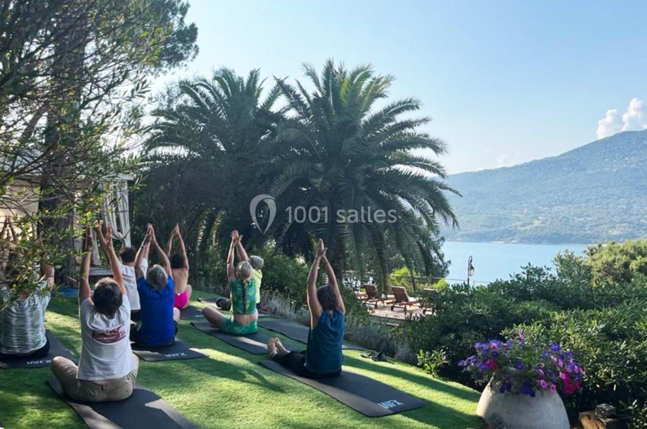 Personnes pratiquant le yoga en plein air sur une pelouse, entourées de végétation avec vue sur la mer et les montagnes.