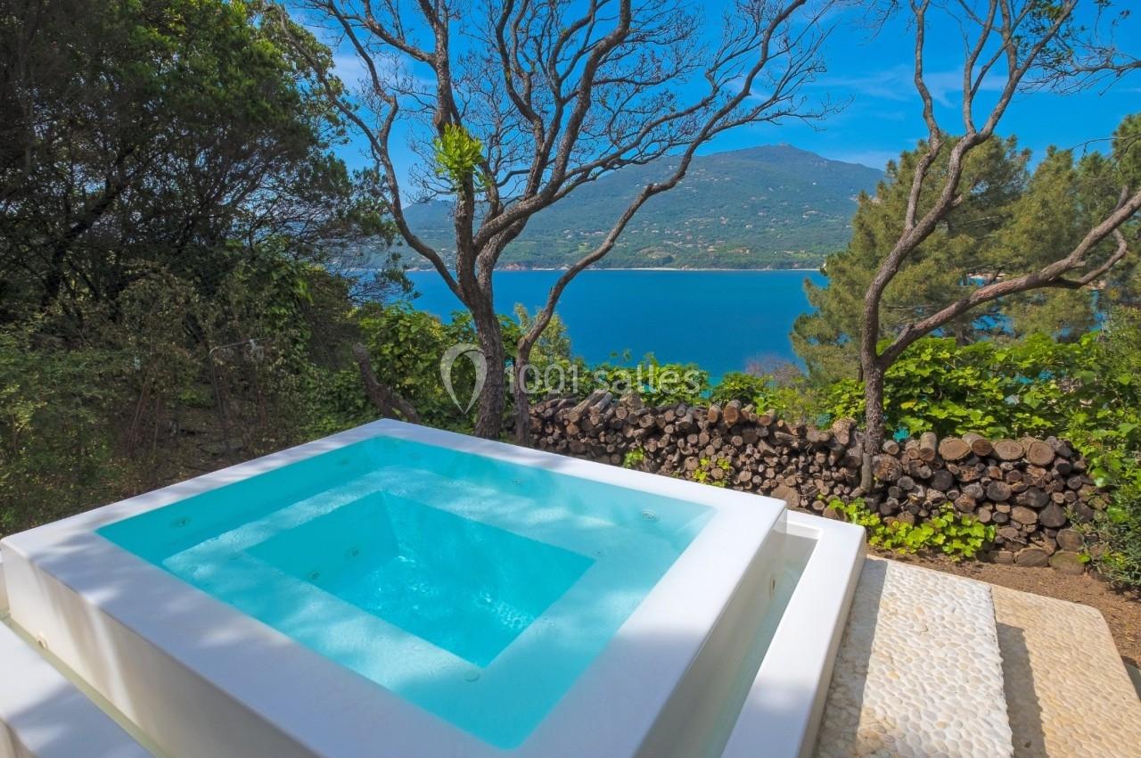 Jacuzzi extérieur blanc entouré de végétation avec vue sur un lac et des montagnes sous un ciel bleu.