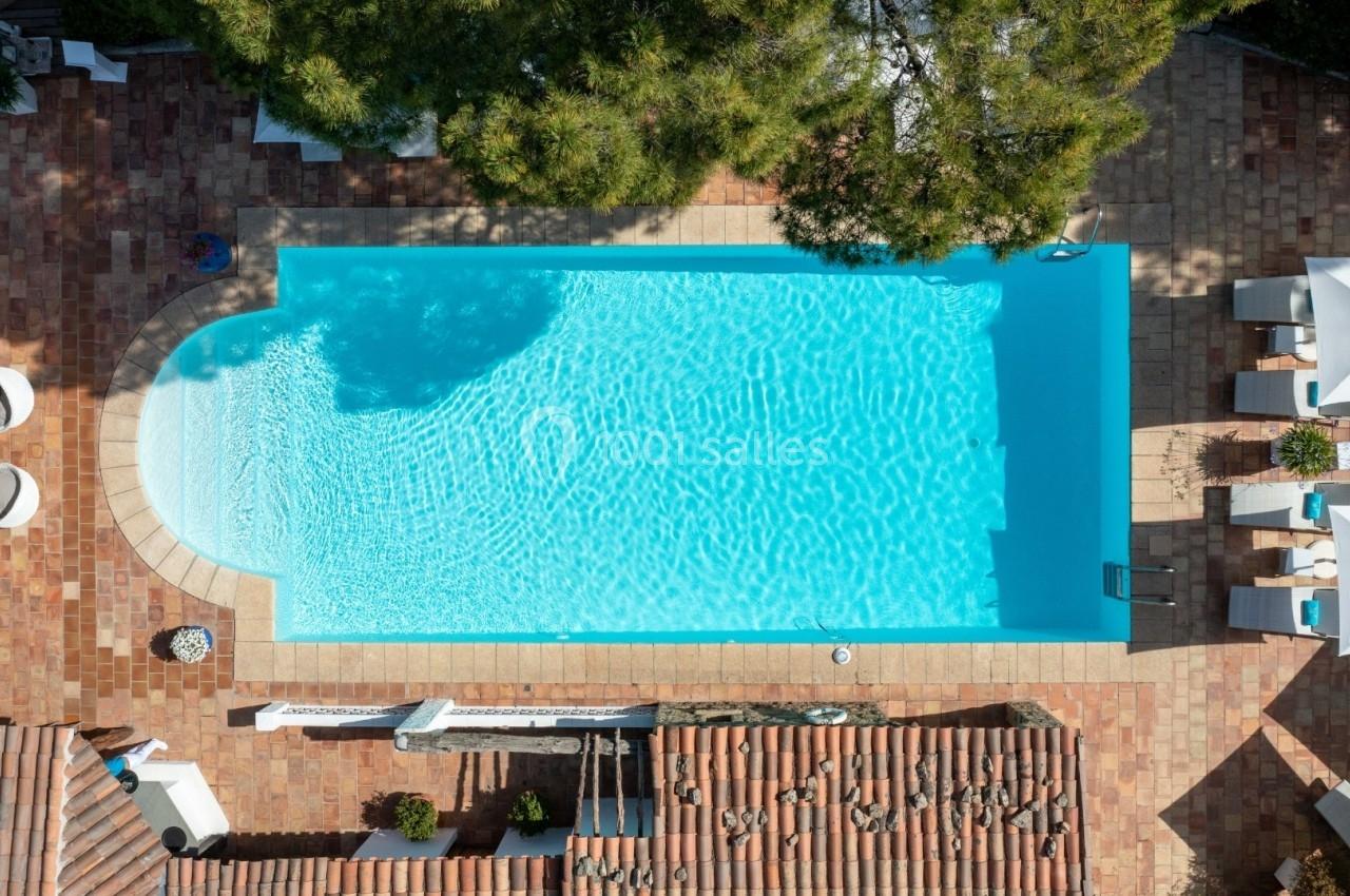 Vue aérienne d'une piscine rectangulaire entourée de terrasses en pierre, de chaises longues et de parasols blancs.