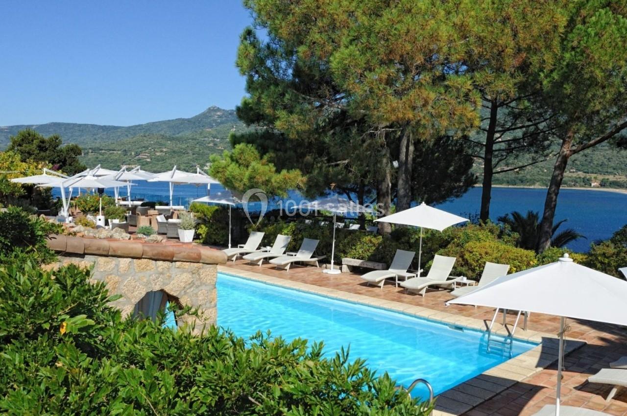 Piscine extérieure entourée de chaises longues et parasols, avec vue sur la mer et des collines boisées en arrière-plan.