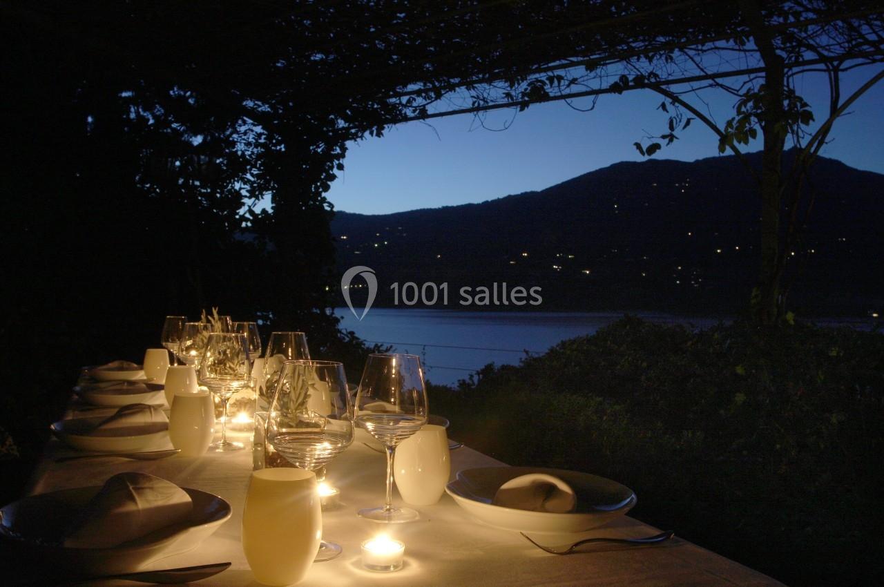 Table dressée avec des bougies et des verres, sous une pergola au crépuscule, avec vue sur un lac et des collines.