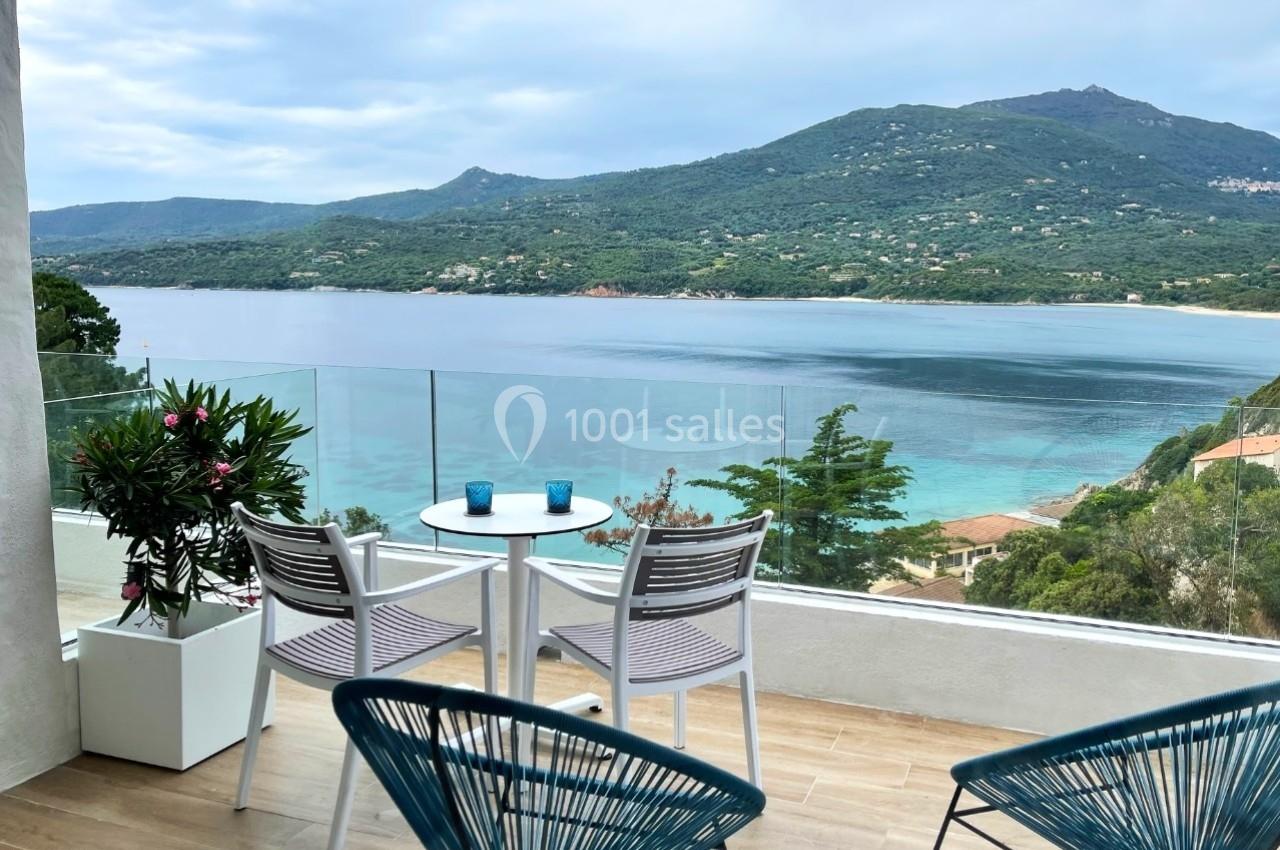 Vue sur une terrasse avec table et chaises, donnant sur une baie entourée de collines verdoyantes et d'eau turquoise.