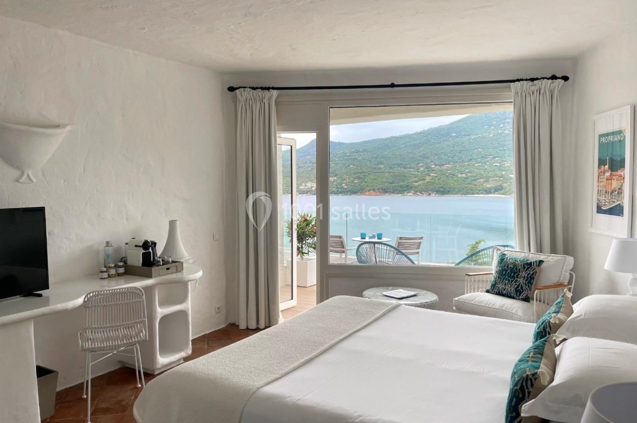 Chambre lumineuse avec lit double, bureau, fauteuil et vue sur une terrasse donnant sur la mer et les collines.