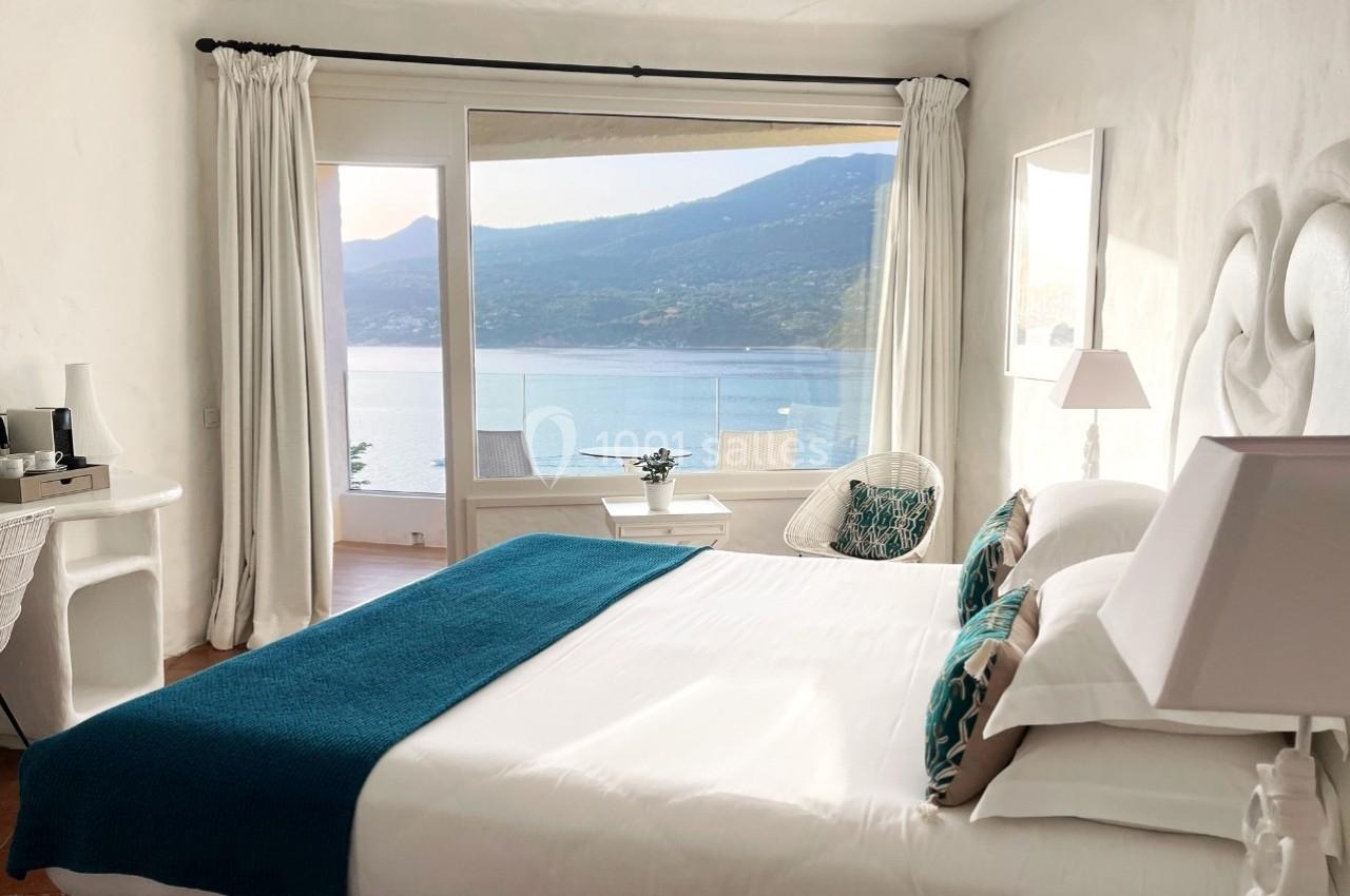 Chambre lumineuse avec lit double, vue sur un lac et montagnes à travers une grande baie vitrée.