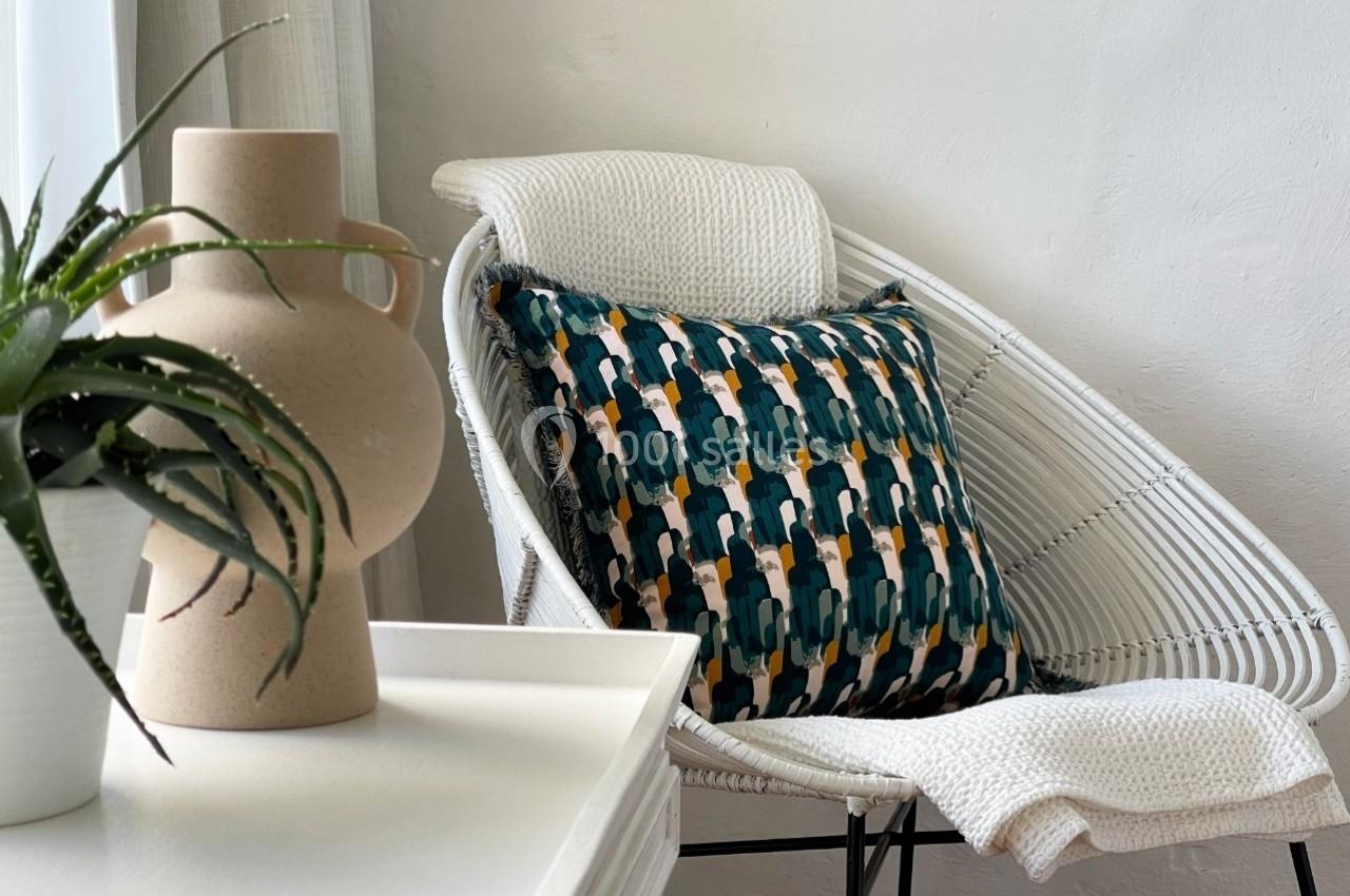 Fauteuil en rotin blanc avec un coussin à motifs géométriques colorés, près d'une table et d'une plante en pot.