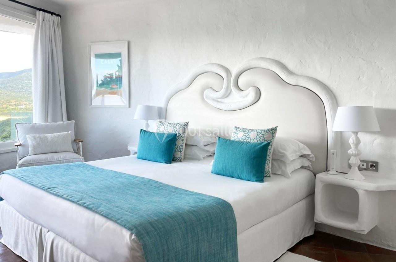 Chambre lumineuse avec lit double, tête de lit blanche sculptée, coussins bleus et vue sur un paysage extérieur.