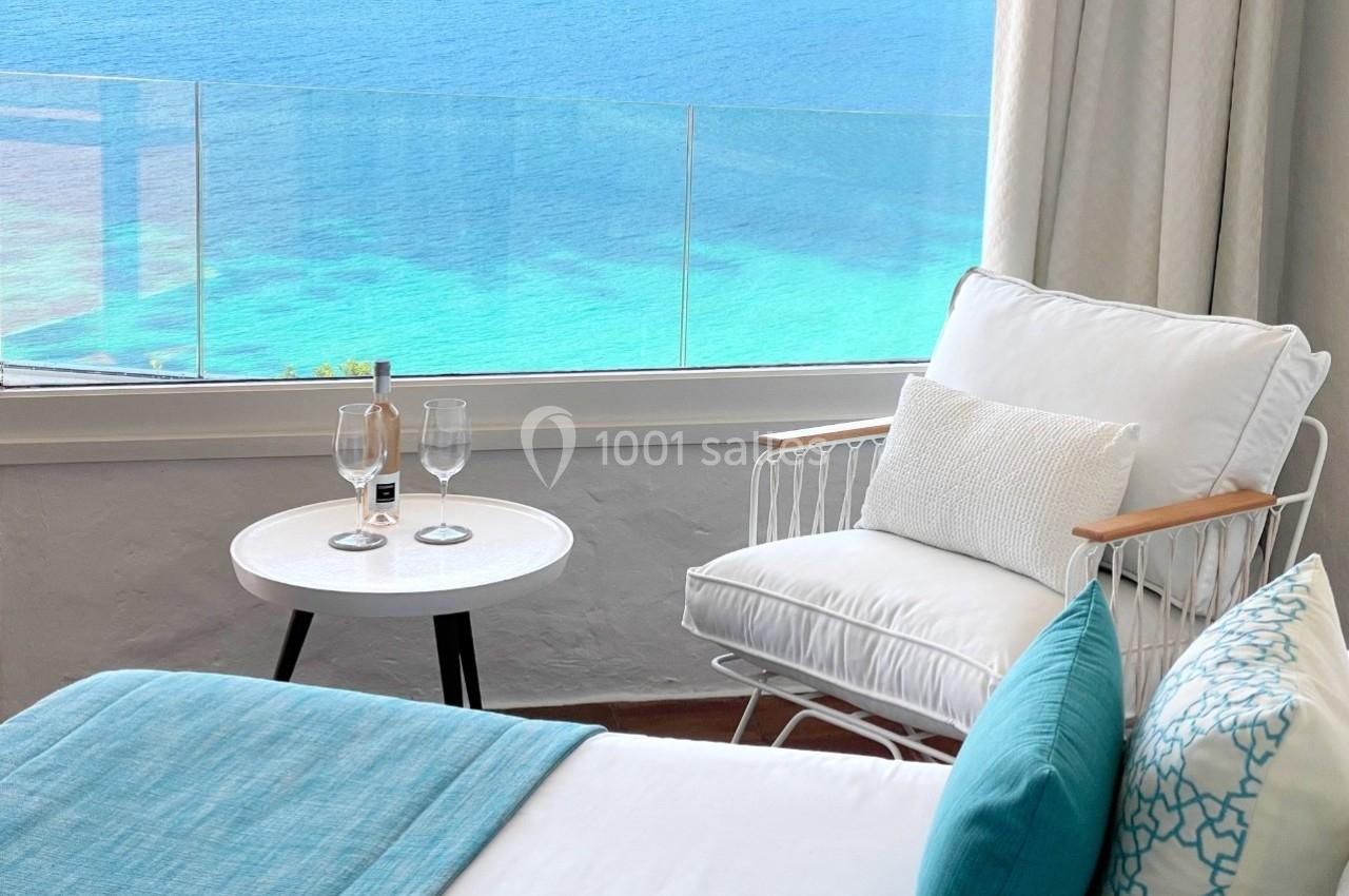 Vue d'une chambre lumineuse avec fauteuil, table basse, lit et grande fenêtre donnant sur la mer turquoise.