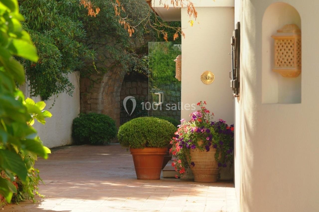 Allée pavée bordée de plantes en pot, menant à une arche en pierre entourée de végétation.