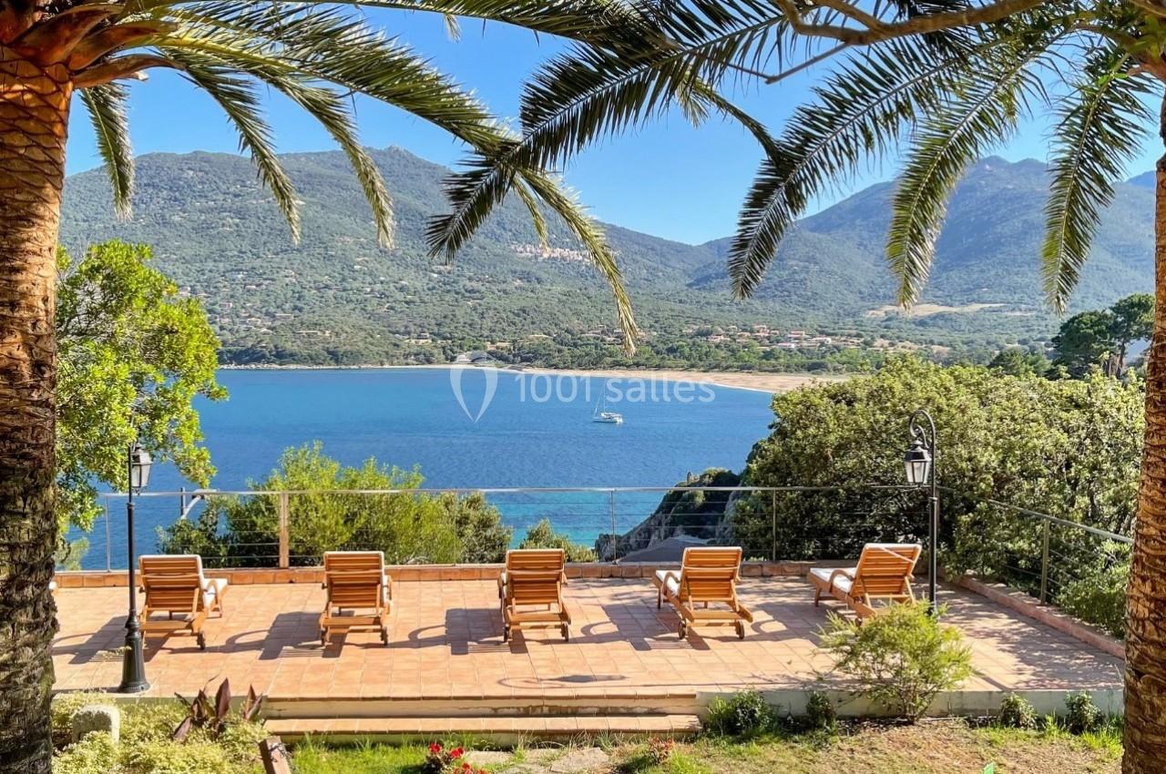 Terrasse avec chaises longues surplombant une baie entourée de montagnes, sous des palmiers.