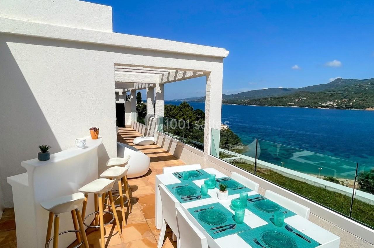 Terrasse ensoleillée avec table dressée, tabourets, vue sur la mer et collines sous un ciel bleu dégagé.
