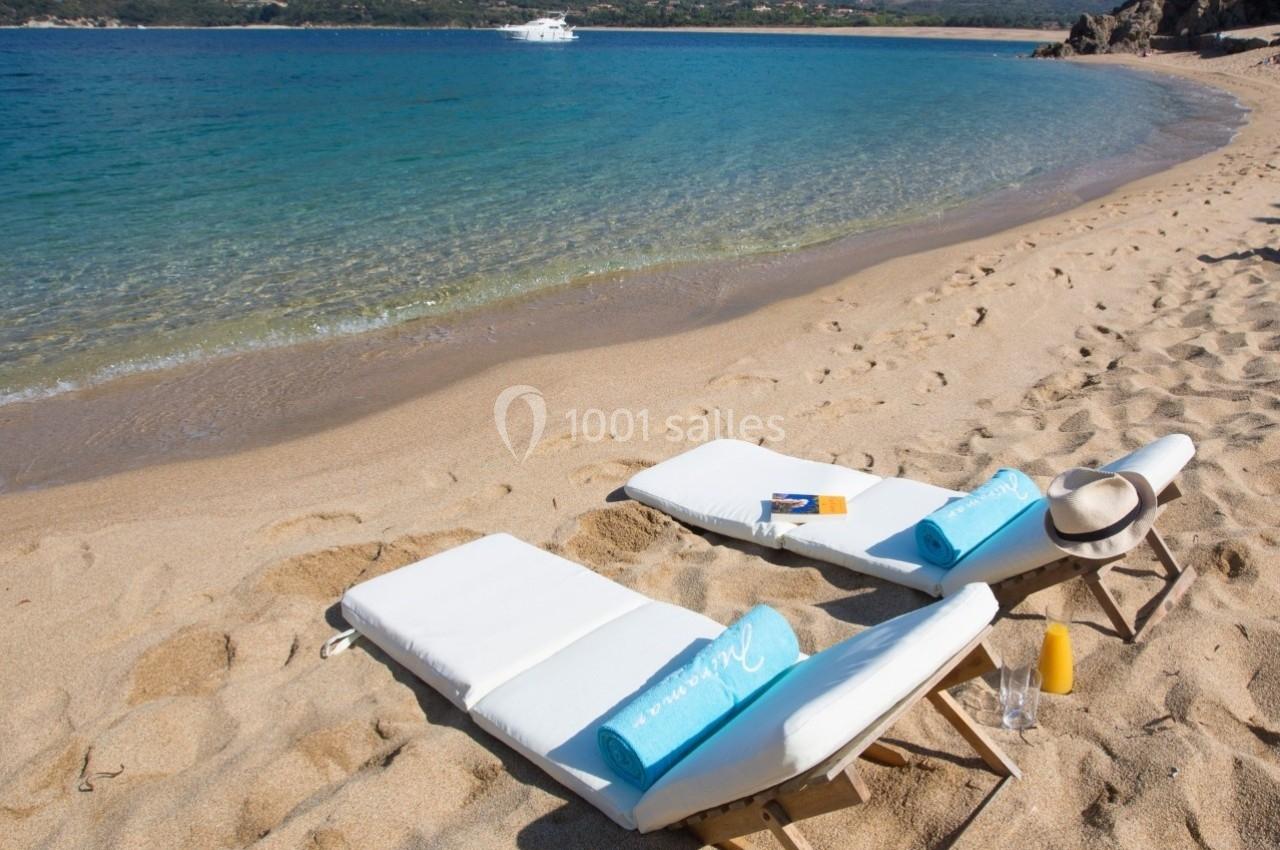 Deux chaises longues avec serviettes bleues sur une plage de sable, face à une mer calme et des collines en arrière-plan.