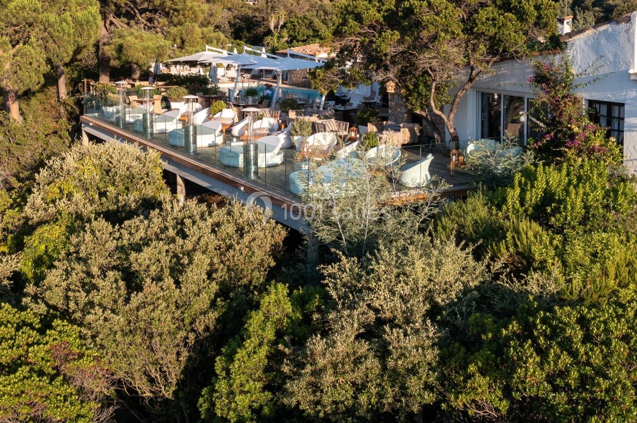 Terrasse en bois surélevée avec tables et chaises, entourée de végétation dense et surplombant un paysage naturel.