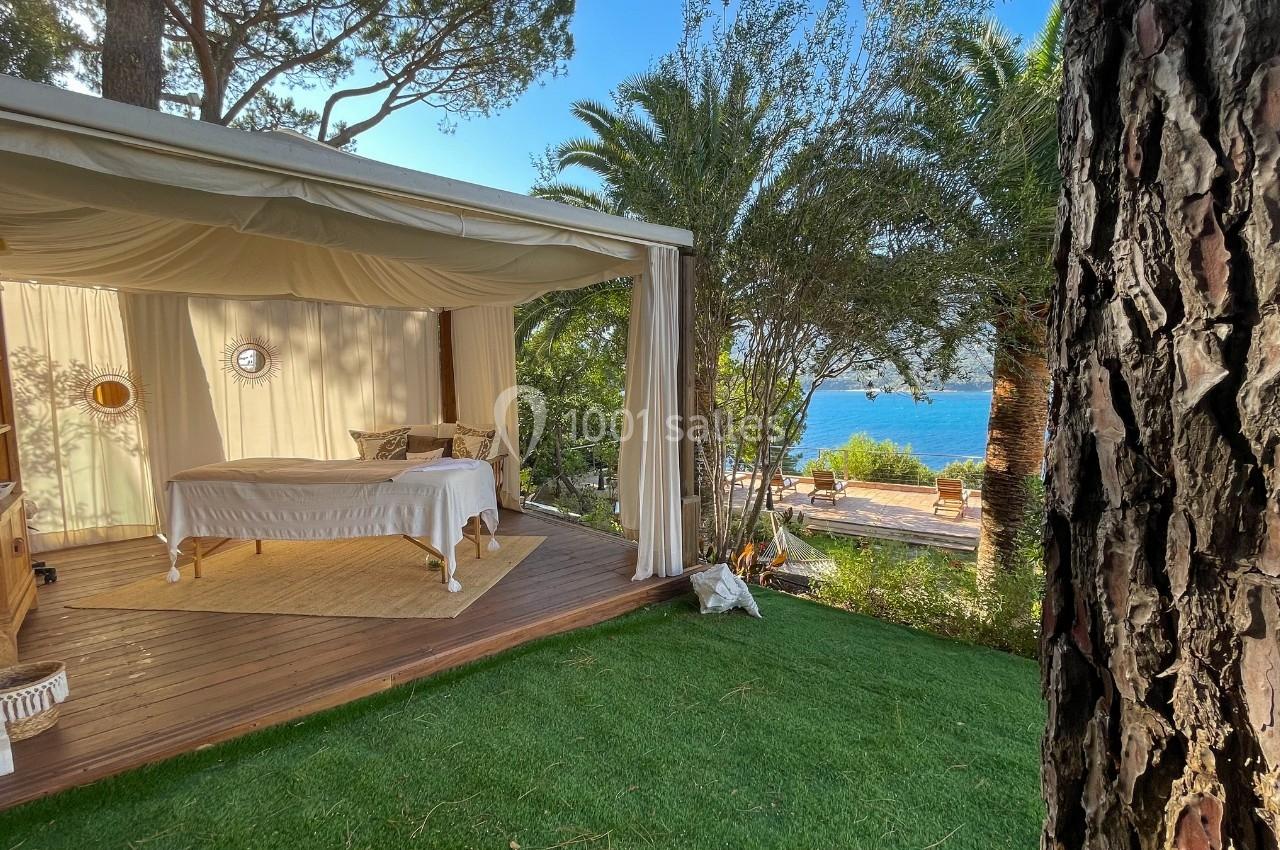 Tente ouverte avec lit sur terrasse en bois, entourée de végétation, vue sur la mer en arrière-plan.