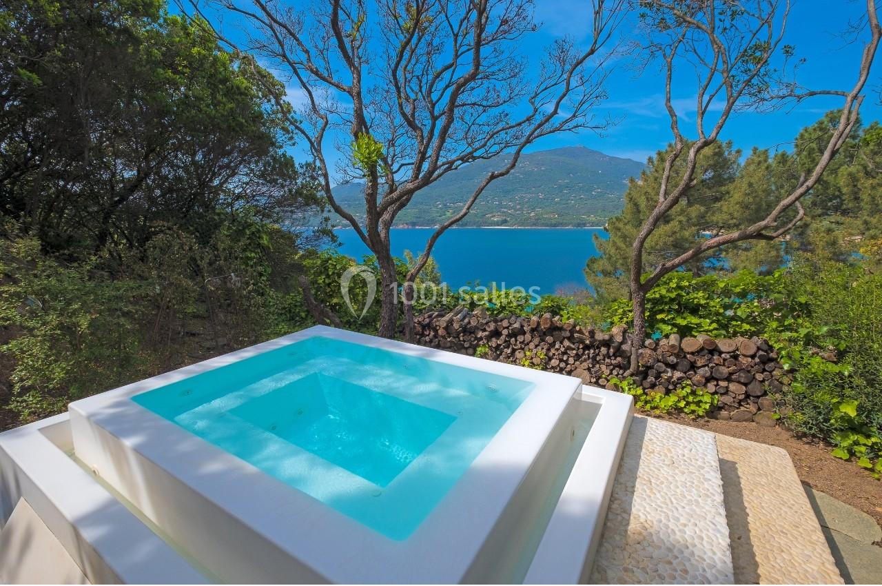 Vue sur un jacuzzi extérieur entouré de végétation, avec un paysage de mer et de montagnes en arrière-plan.