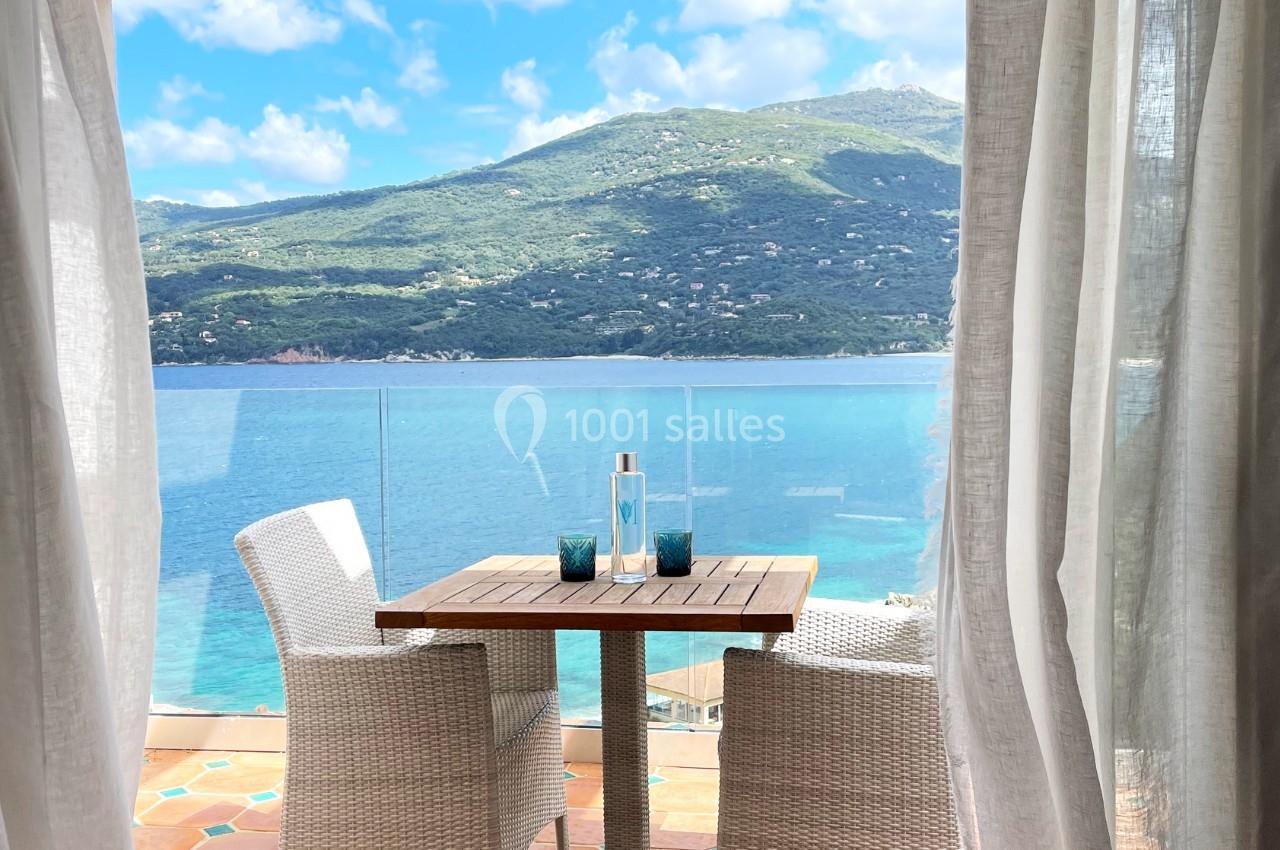 Vue sur une terrasse avec table et chaises en rotin, donnant sur une mer bleue et des collines verdoyantes à l'horizon.