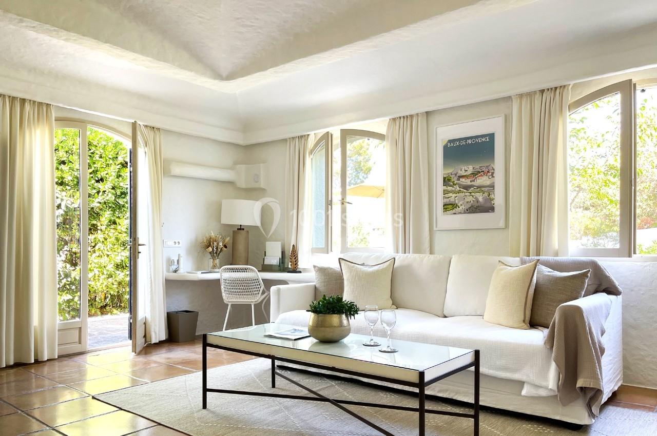 Salon lumineux avec canapé blanc, table basse, bureau près de la fenêtre et vue sur un jardin verdoyant.