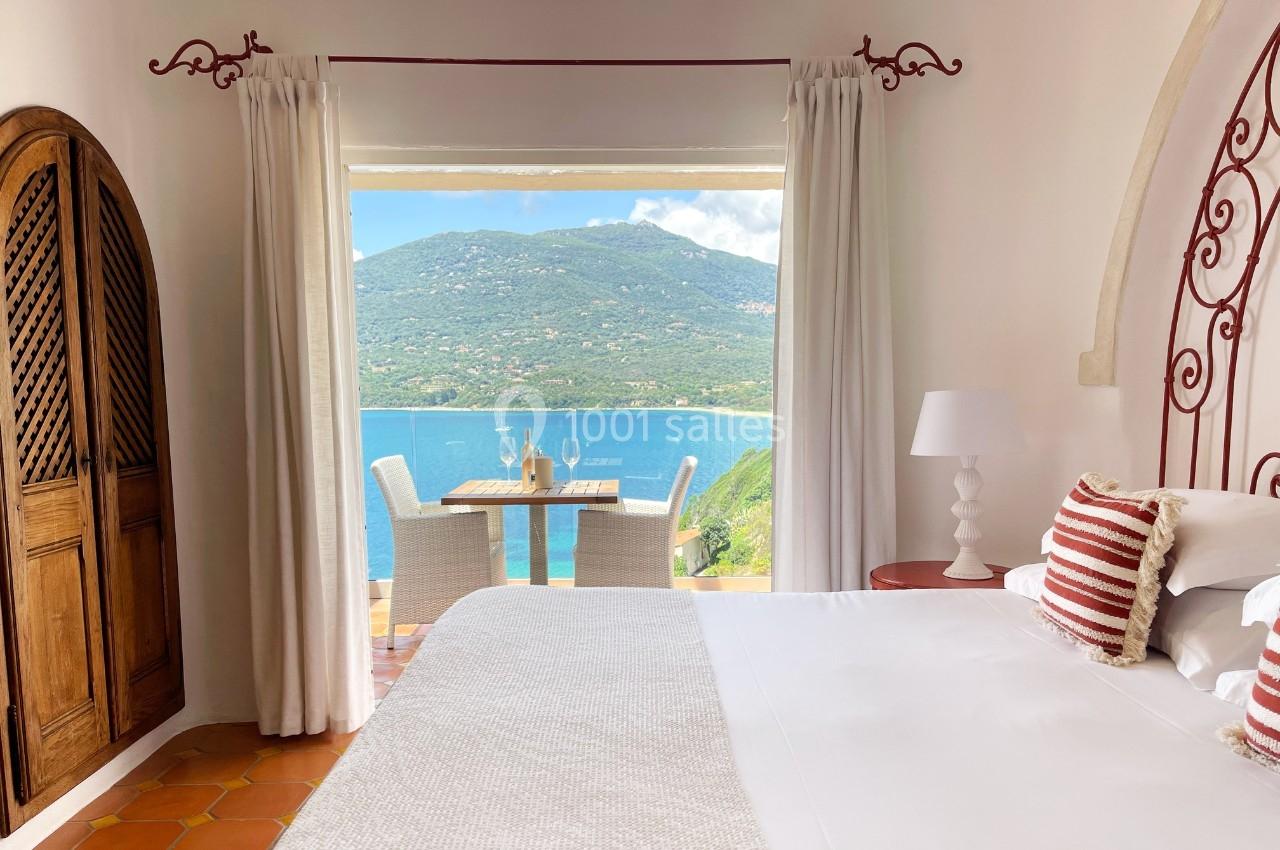 Chambre lumineuse avec lit, rideaux ouverts sur une terrasse offrant une vue sur la mer et des montagnes verdoyantes.