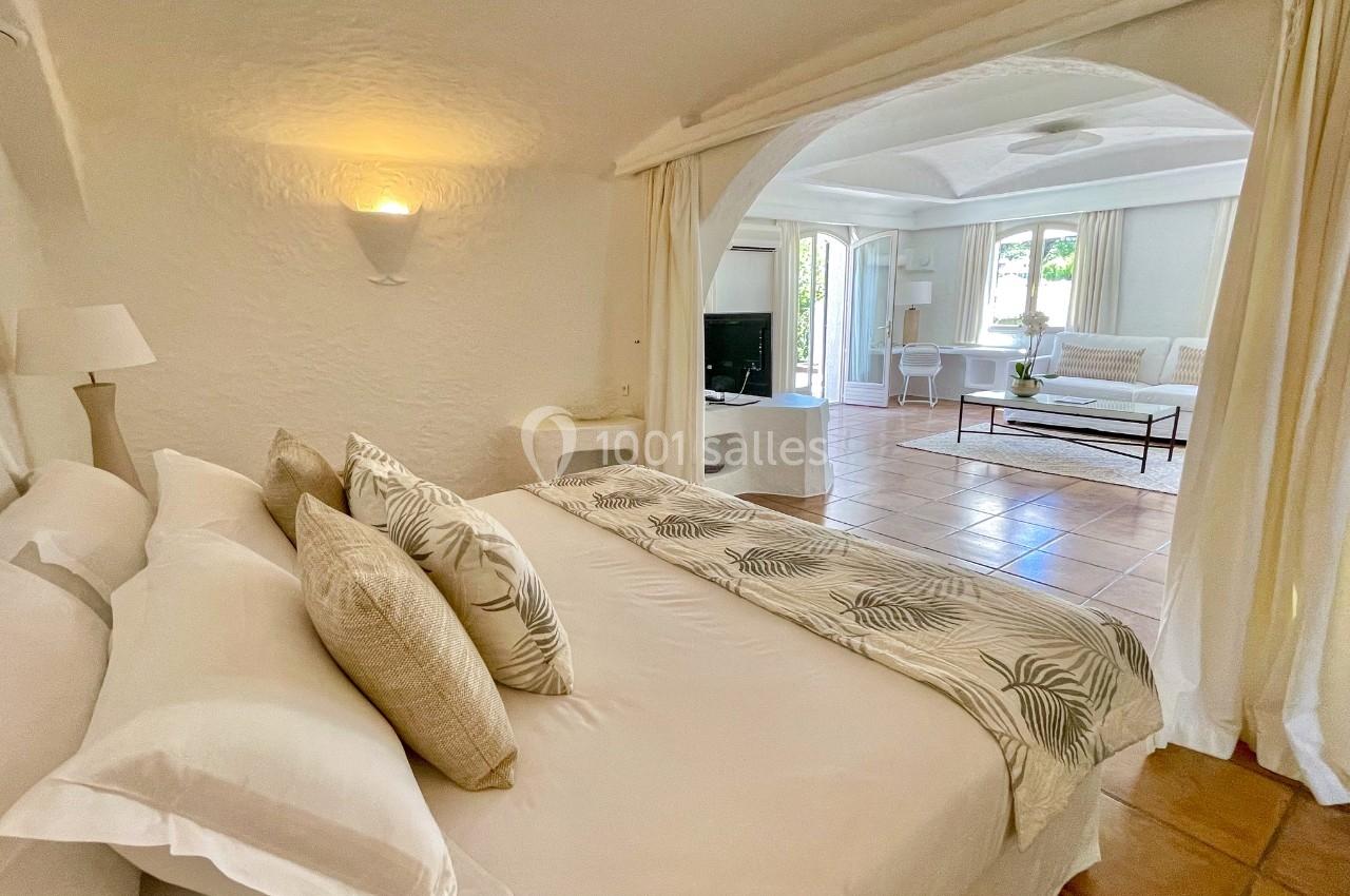 Chambre lumineuse avec lit double, coussins décoratifs et vue sur un salon spacieux à travers une arche.