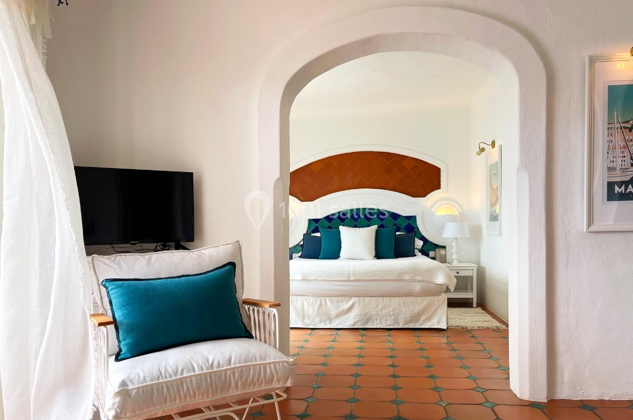 Chambre lumineuse avec lit double, coussins colorés, fauteuil, télévision et sol en carrelage terracotta.