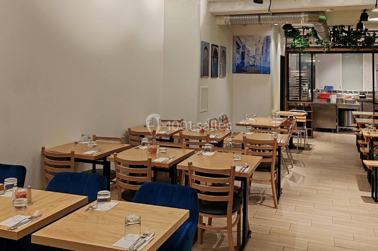 Salle de restaurant lumineuse avec des tables en bois, des chaises en bois et un espace cuisine visible en arrière-plan.