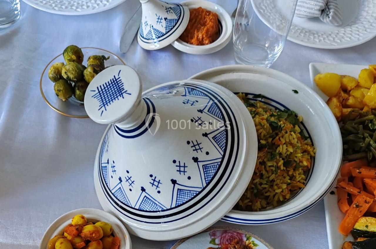 Plat de tajine en céramique blanche décorée, entouré de légumes variés et de condiments sur une nappe blanche.