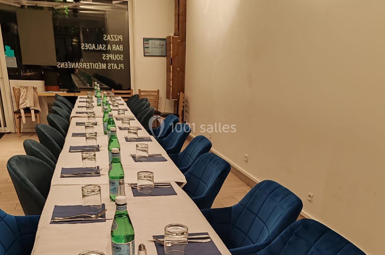 Salle de restaurant avec une longue table dressée, nappes blanches, chaises bleues et bouteilles d'eau minérale.