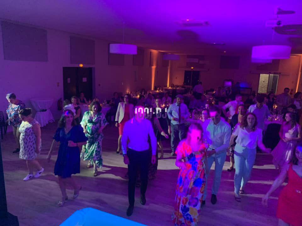 Des personnes dansent dans une salle éclairée par des lumières colorées, avec des tables dressées en arrière-plan.