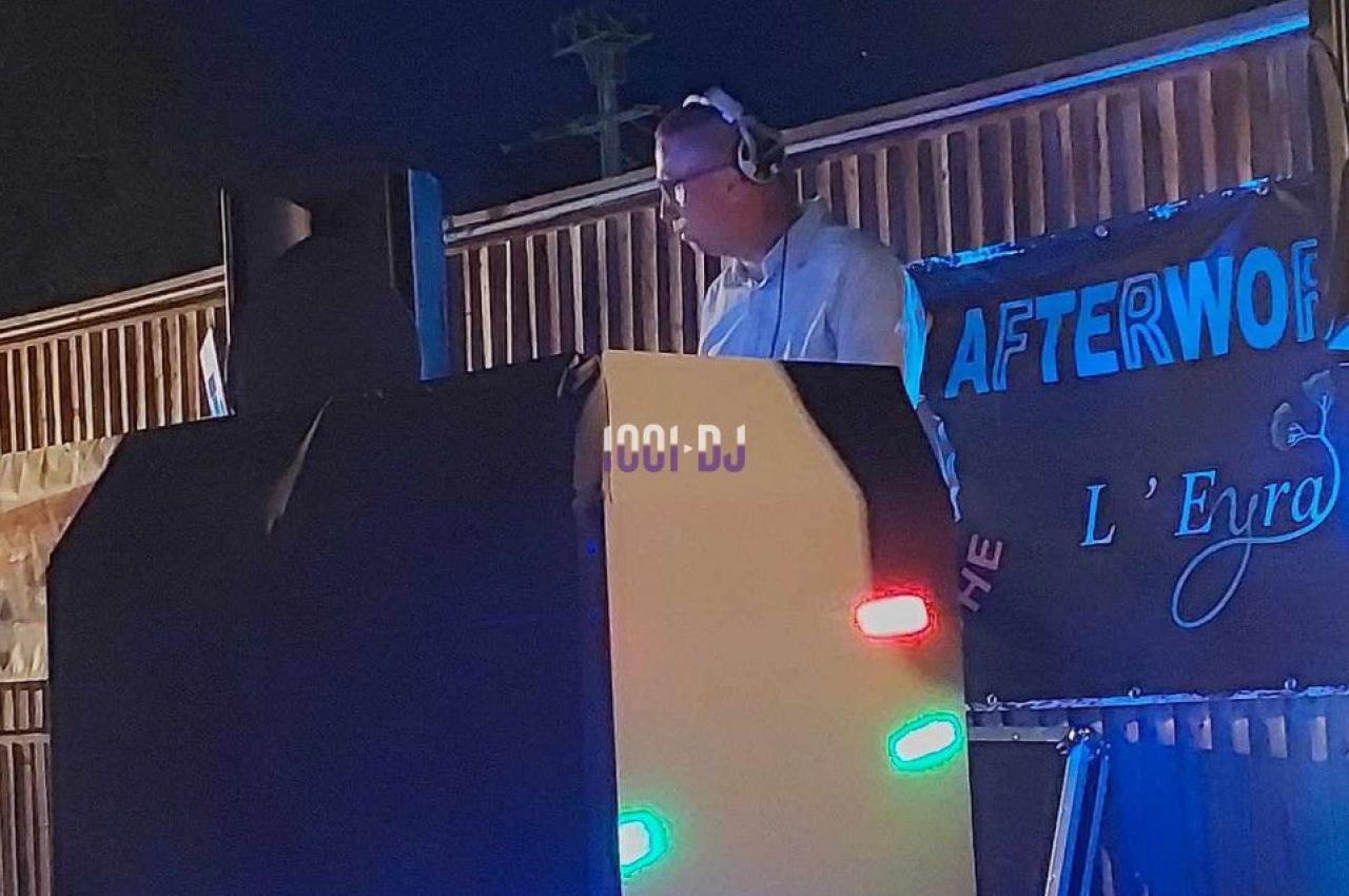 Un DJ mixe en extérieur de nuit, éclairé par des lumières colorées devant une bannière.