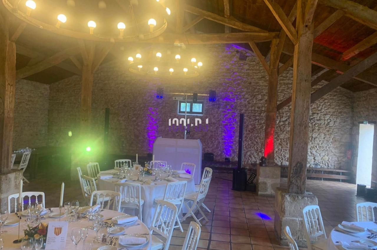 Salle de réception en pierre avec tables dressées, éclairage coloré et poutres apparentes en bois.
