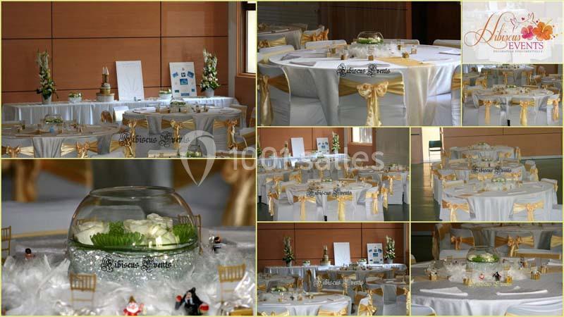 Salle décorée pour un événement avec des tables rondes habillées de nappes blanches, ornées de rubans dorés et centres de…