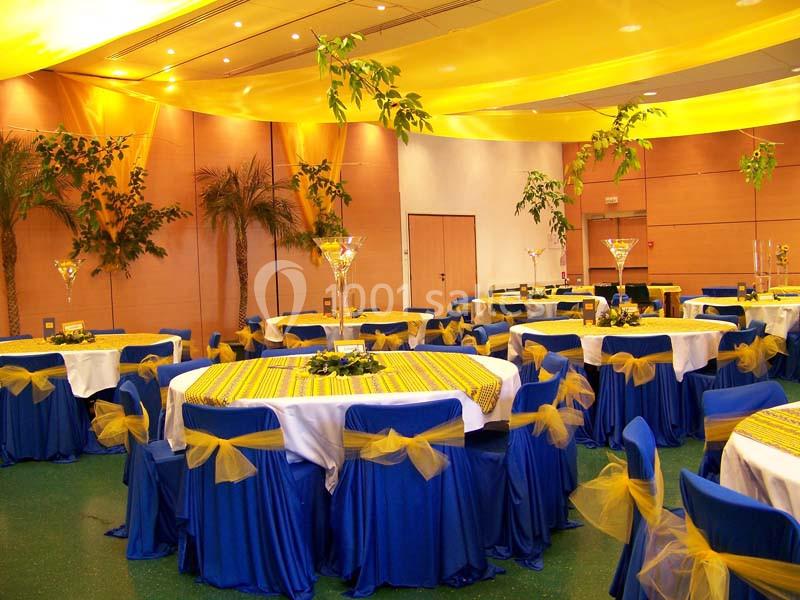 Salle décorée pour un événement avec des tables rondes, nappes jaunes et bleues, chaises ornées de rubans jaunes.
