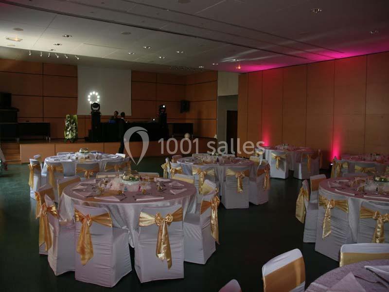 Salle de réception décorée avec des tables rondes, nappes violettes et chaises ornées de nœuds dorés.
