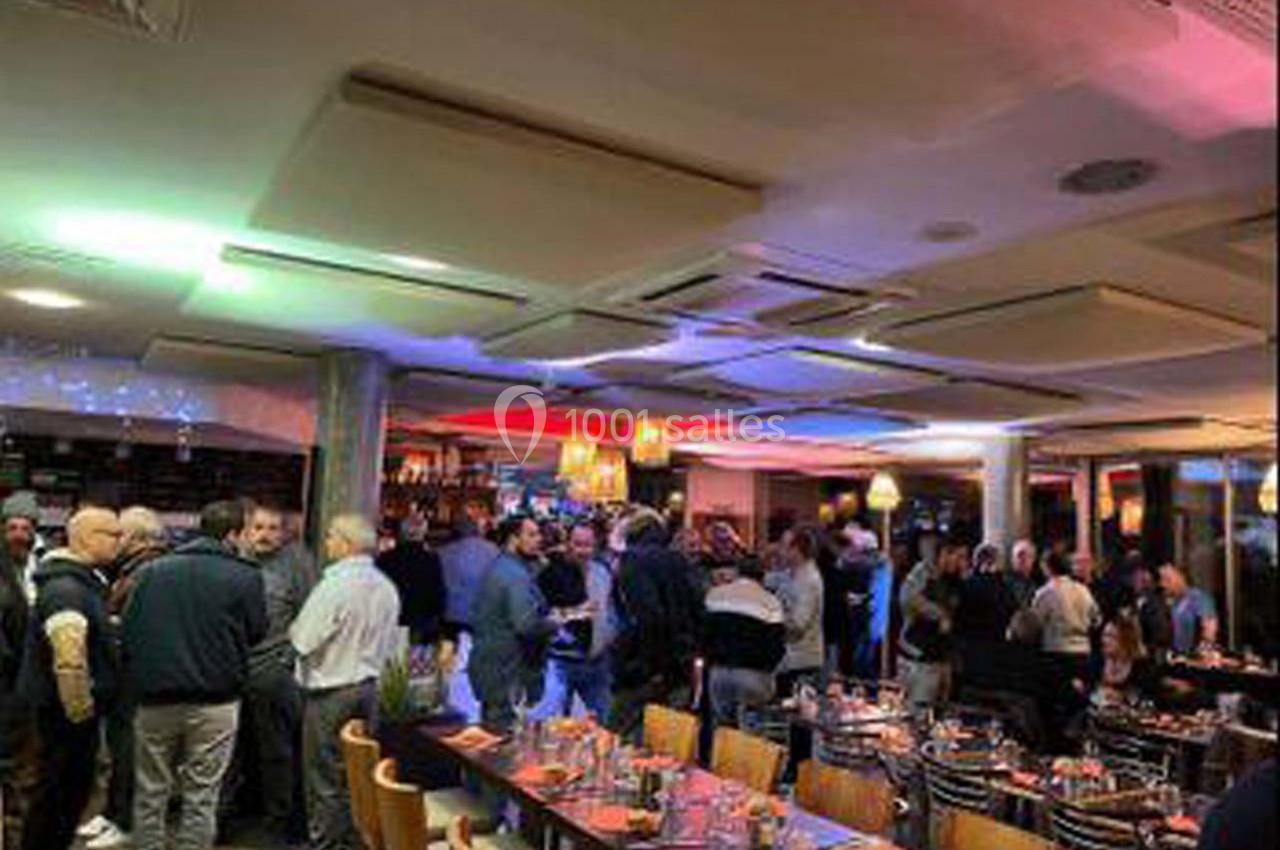 Salle de restaurant animée avec des convives debout près du bar et des tables dressées en premier plan.