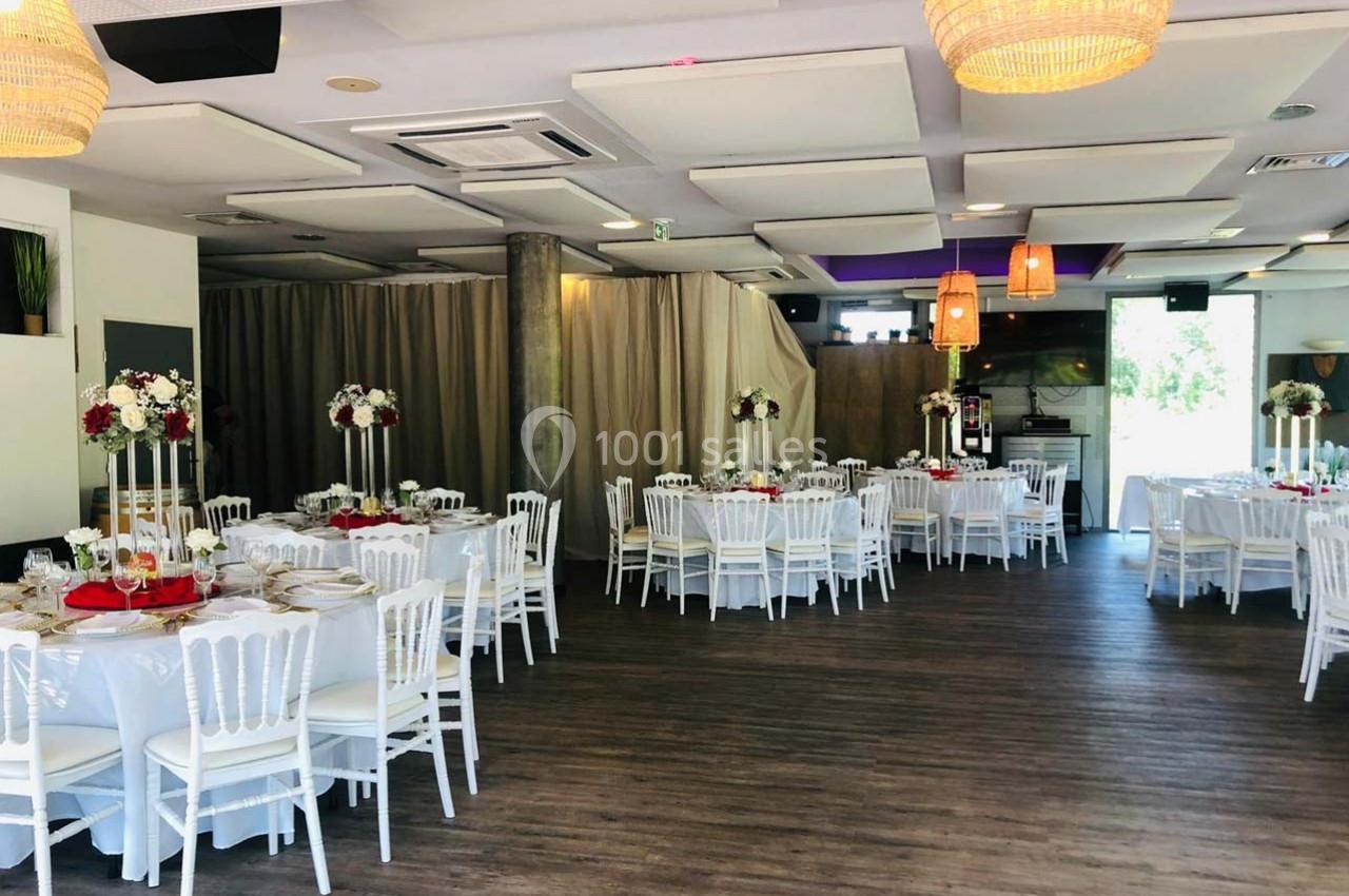 Salle de réception décorée avec des tables rondes, nappes blanches, chaises blanches et arrangements floraux.